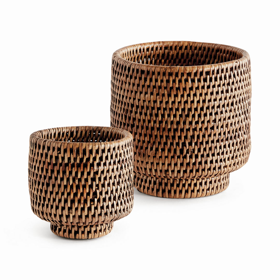 Brown rattan cachepots in two sizes with cylindrical woven sides and small round feet, shown on white - Coastal Transitional