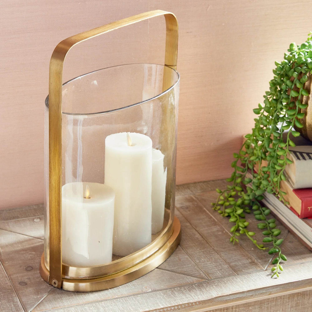 Large oval brass and glass hurricane candle holder with two pillar candles styled on console table with books and greenery