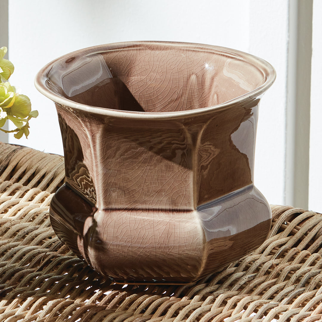 Brown reactive glaze ceramic planter with faceted sides, displayed on a woven surface beside a bright window, Traditional