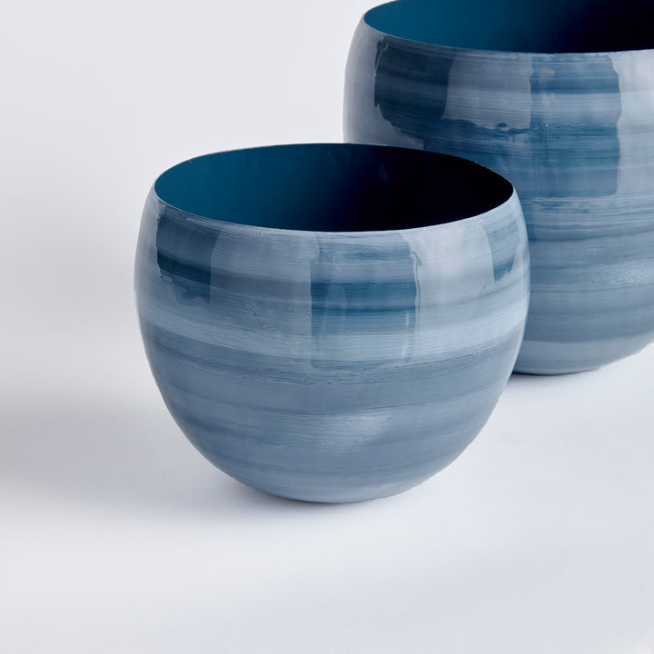 Crafted from lightweight enameled iron and meticulously hand-painted by experts, the Andrey Cache Planter Pots boast a refined, horizontal swirl design in a coastal-inspired blue gray palette. These cachepots serve as both functional pieces and unique works of art, adding a touch of modern sophistication to any space.