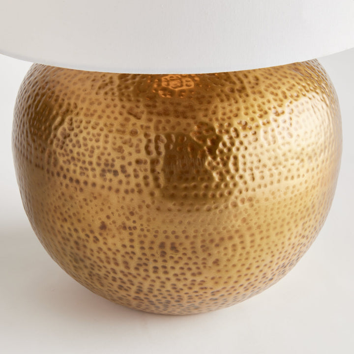 Crafted with a round base in warm brass and an impressive scale, the Tara Gold &amp; White Table Lamp serves as a handsome anchor for any room. Its clean white linen drum shade adds a soft touch and complements the bold design. Made from hammered iron, this one-of-a-kind lamp is sure to catch the eye and become a stunning focal point in your space.