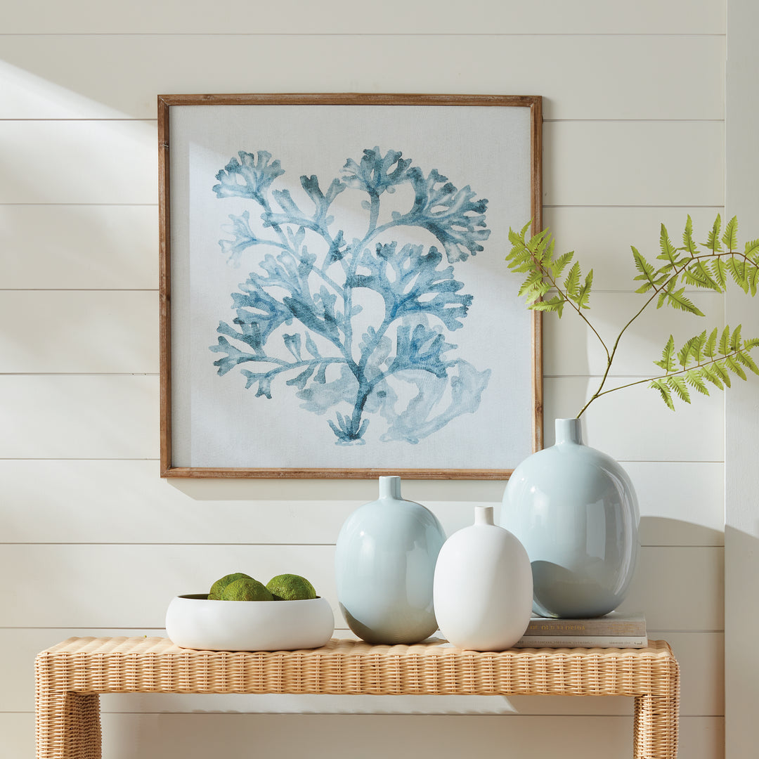 Blue coral framed print displayed above a woven console with pale vases and green stems, in a square wood frame. Coastal