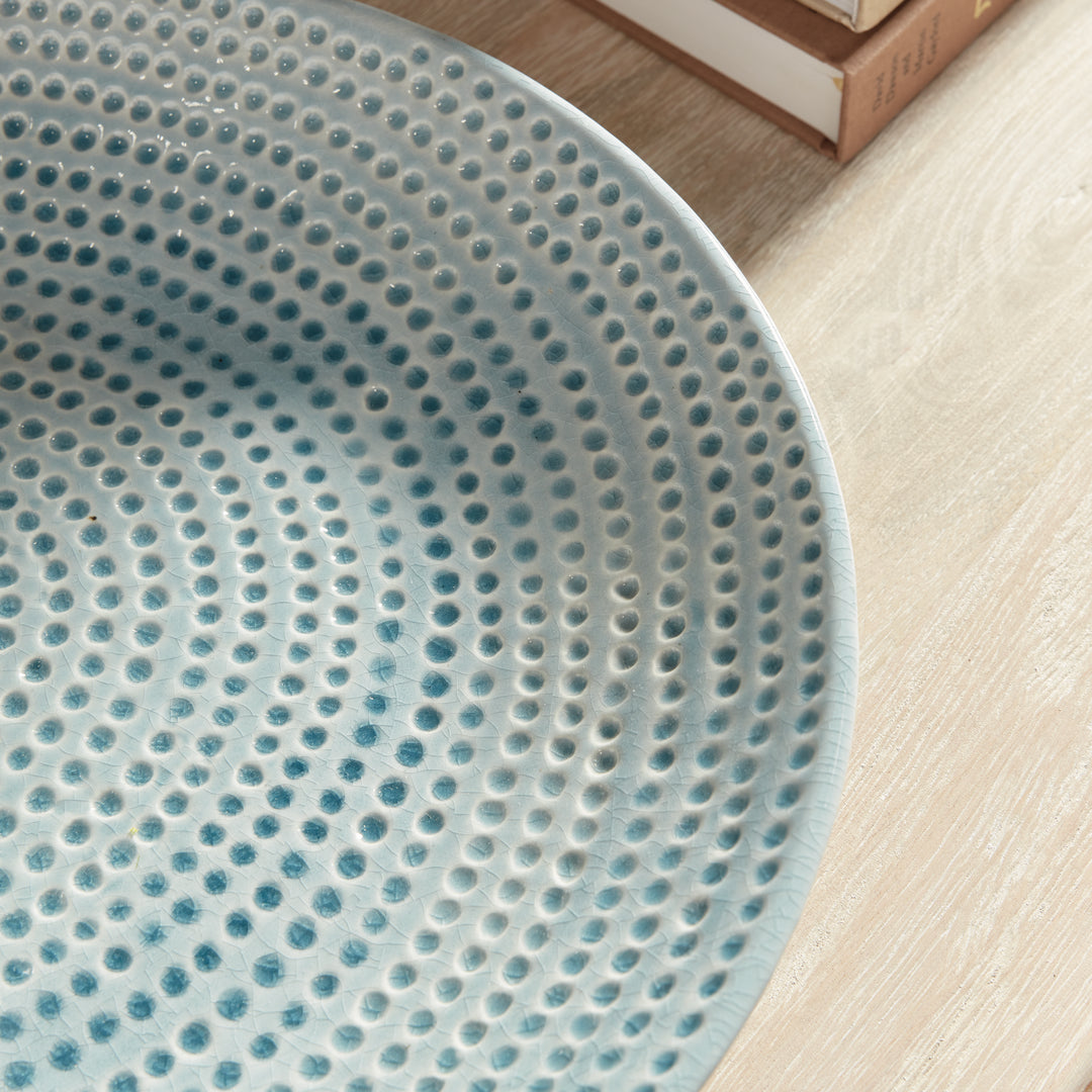 Angled close view of Carys blue decorative plate with rows of glazed dots and a shallow rim beside books on a table. Coastal