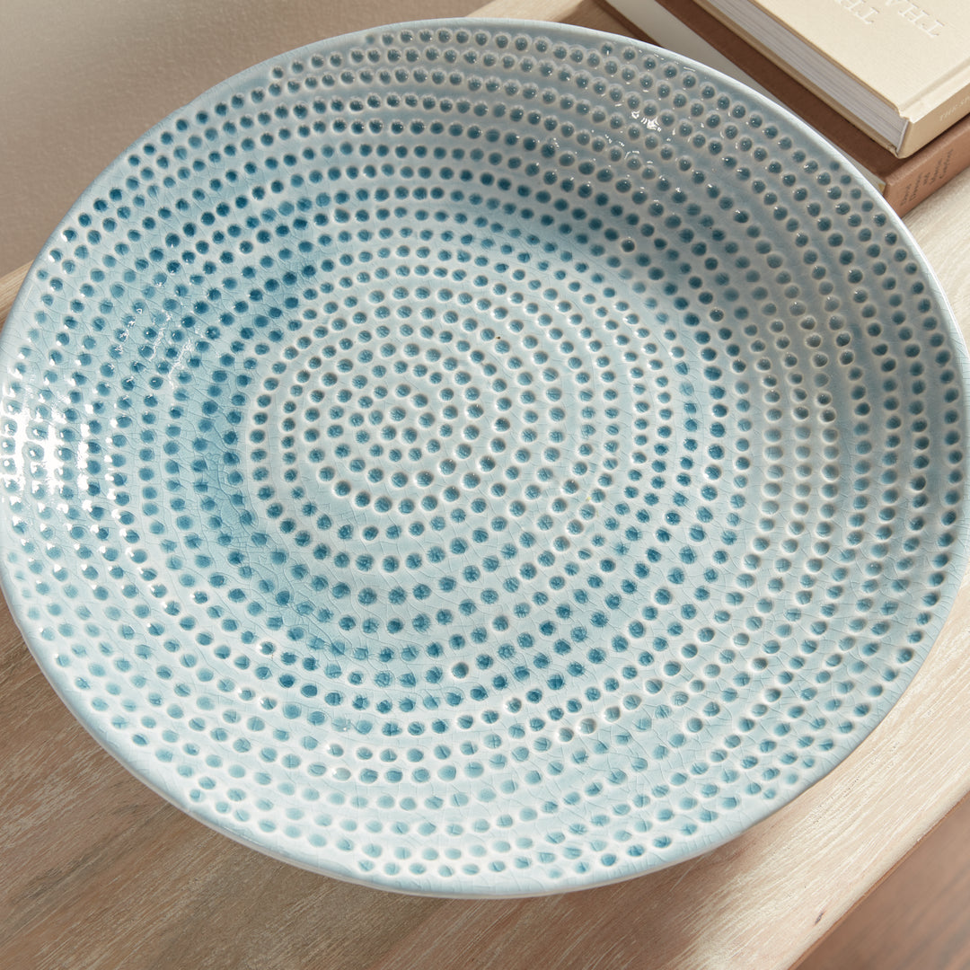 Overhead detail of Carys blue decorative plate showing the off-center dotted texture beside books on a wood console. Coastal