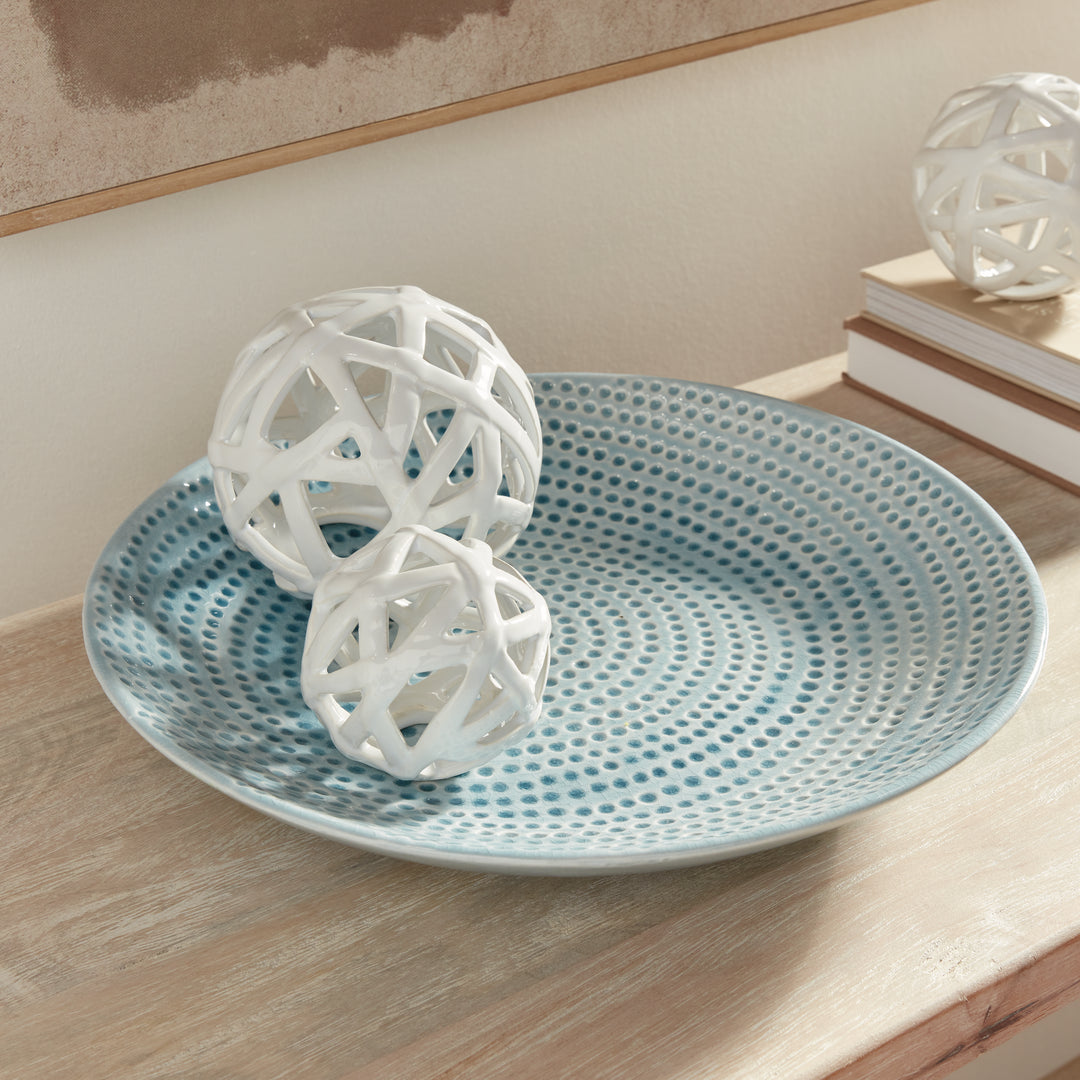 Carys blue dotted decorative plate holding two white openwork spheres on a wood console beside books and framed art. Coastal