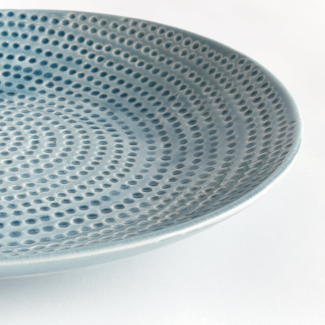 Close view of Carys blue decorative plate showing the curved rim and dotted glazed surface against a pale background. Coastal