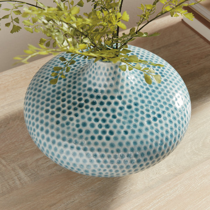 Angled detail of Carys low blue dimpled vase with leafy stems above its broad glazed shoulders on a wood tabletop. Coastal