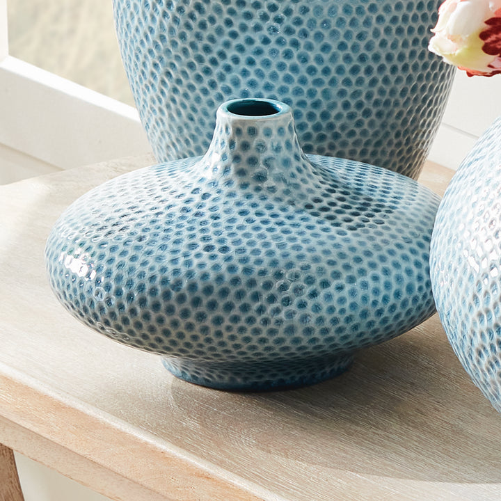 Carys low narrow-neck blue vase among coordinating dimpled ceramics on a wood tabletop with pale flowers nearby. Coastal