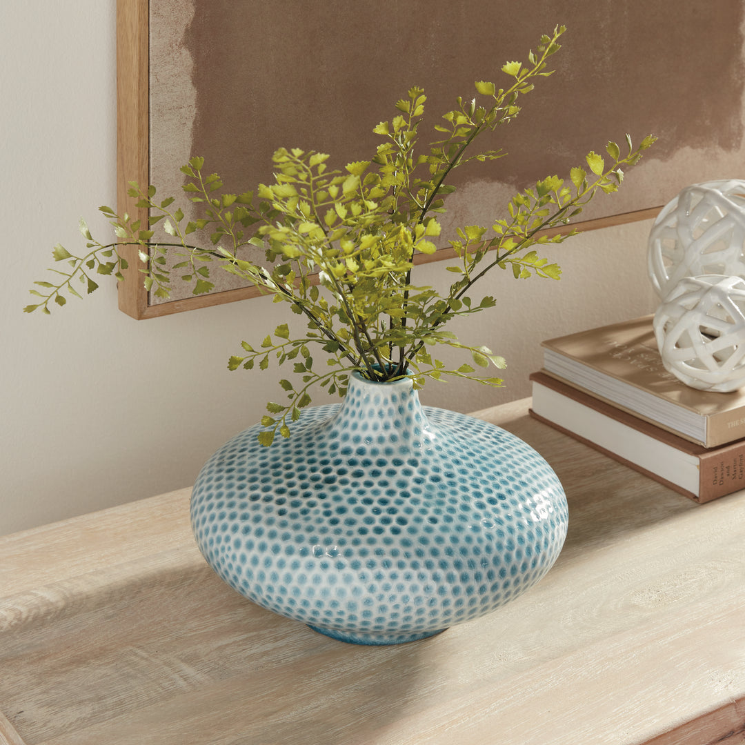 Carys low blue vase with leafy green stems on a wood console beside books and white openwork decorative spheres. Coastal
