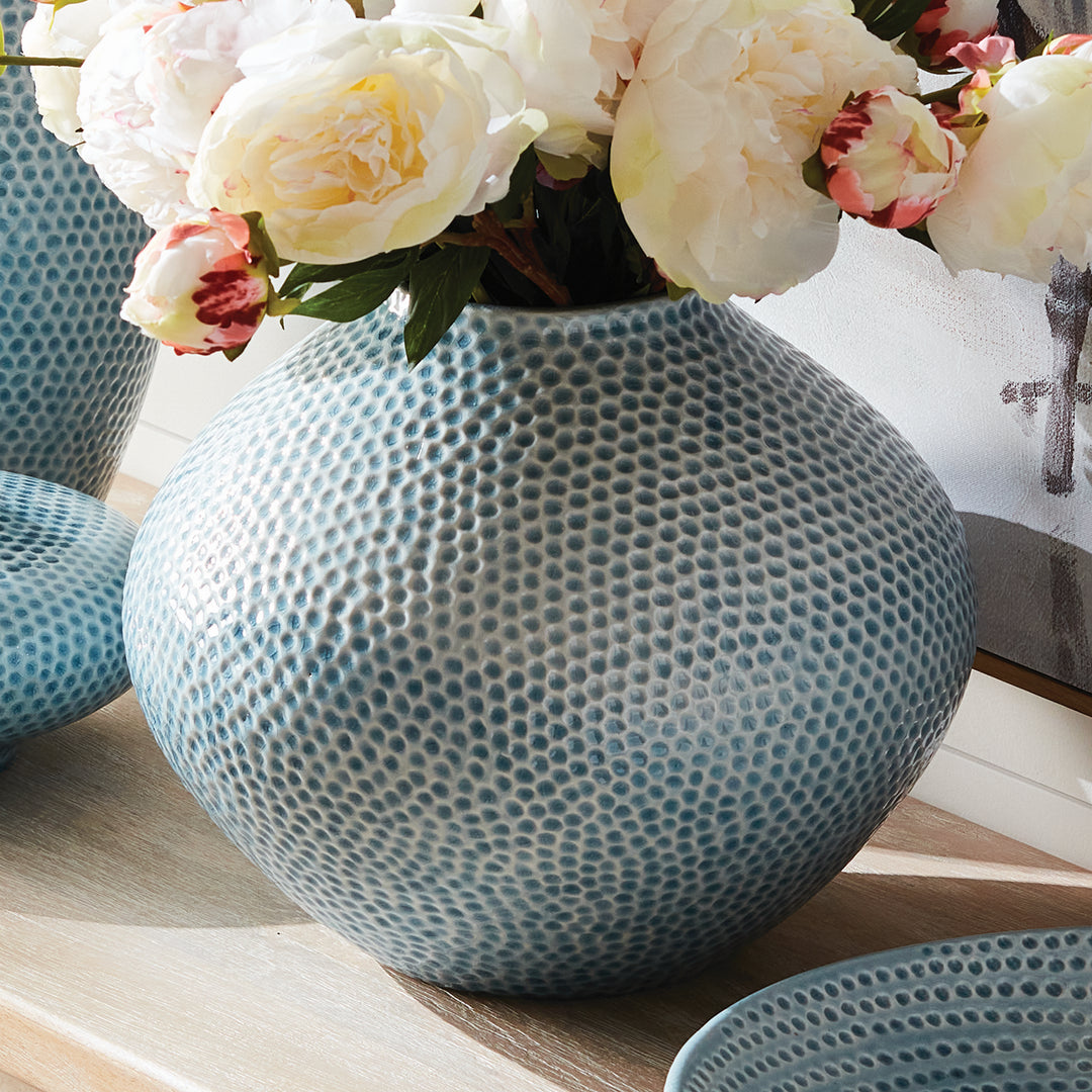 Carys rounded blue vase with pale flowers beside coordinating ceramic pieces on a wood table beneath framed artwork. Coastal