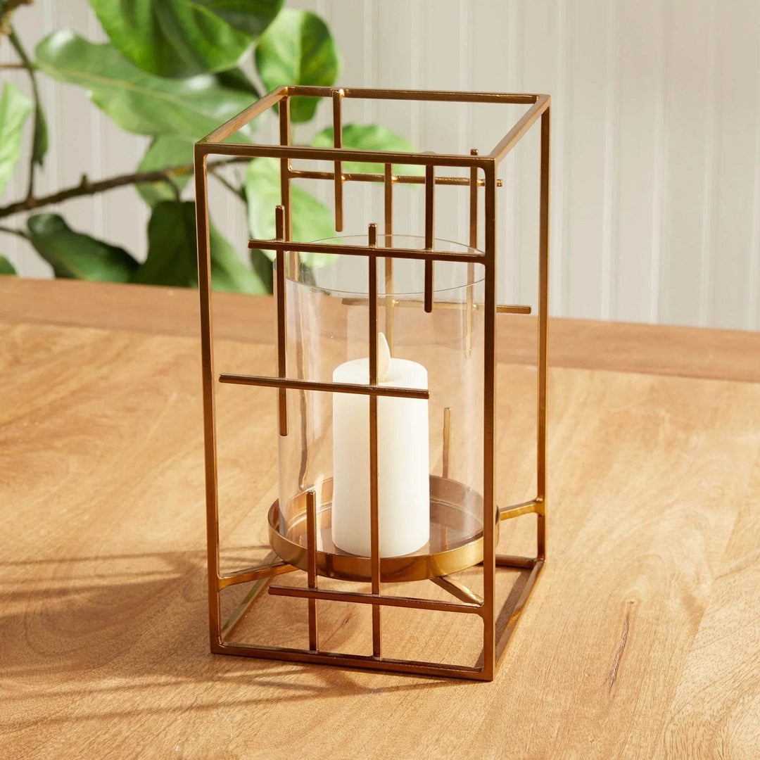 Gold metal hurricane candle holder with glass cylinder, styled on a wood tabletop near leafy green branch – Art Deco Revival

