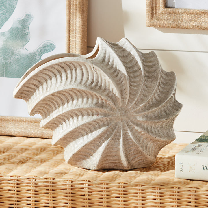 Cream shell vase displayed on woven console beneath framed artwork, showing its spiral form and raised glazed ridges. Coastal