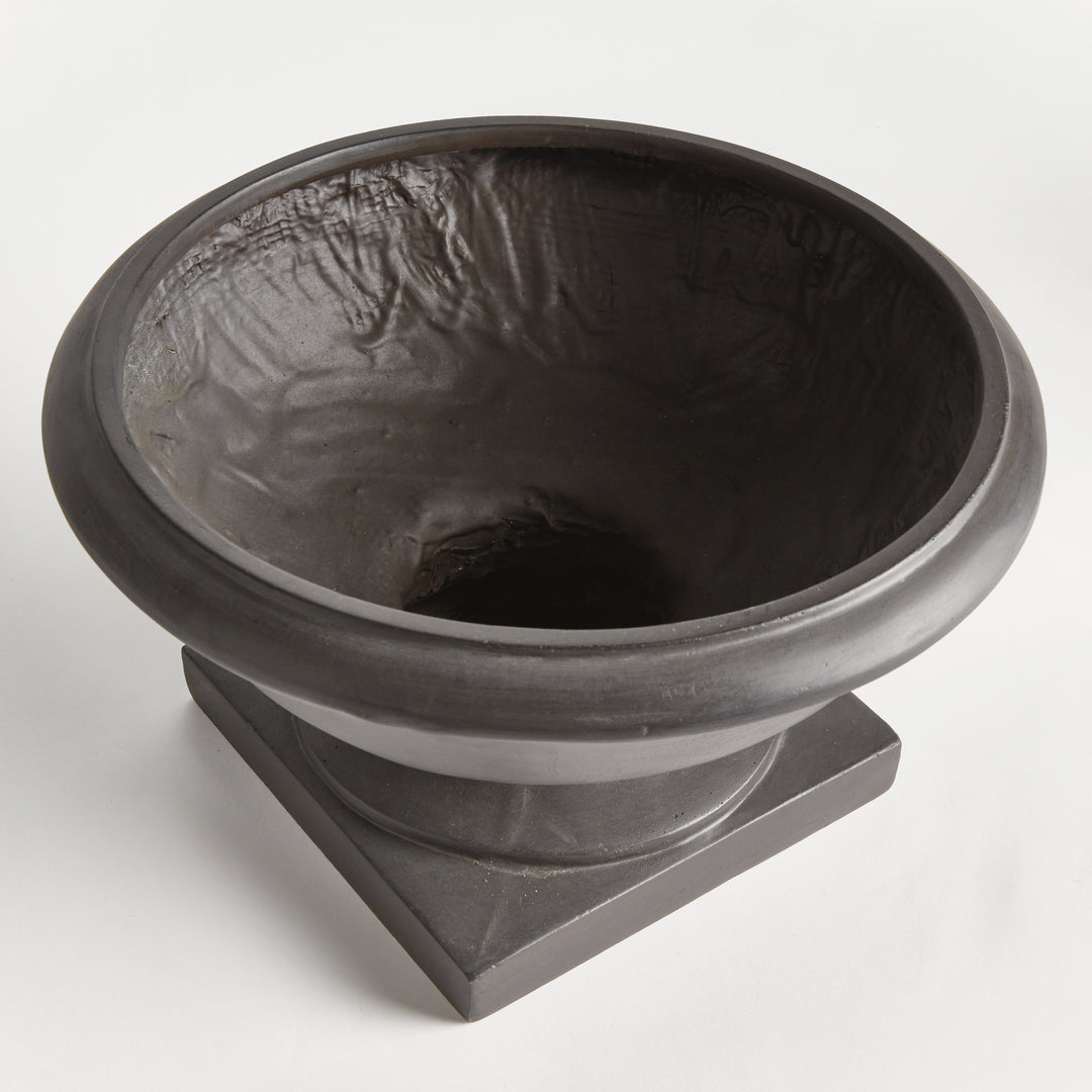 Angled view of an antique black footed bowl planter showing a wide opening, curved basin and square raised base. Transitional