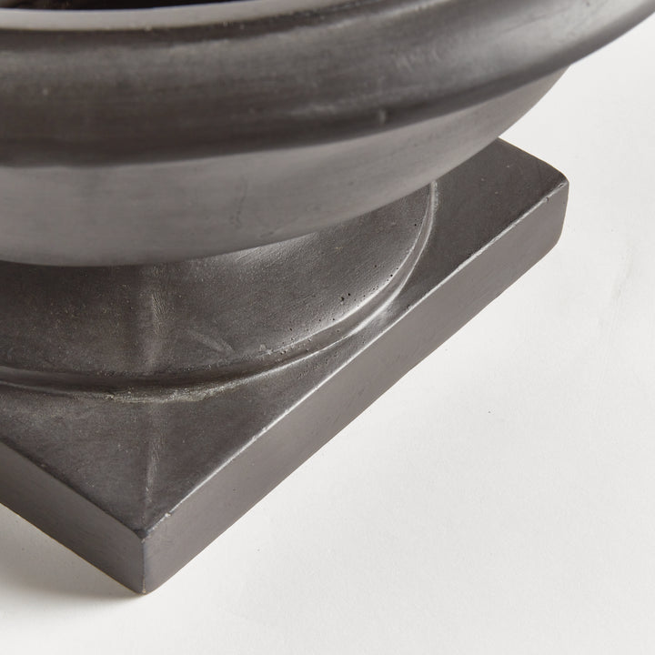 Close detail of an antique black bowl planter showing a curved pedestal foot and layered square composite base. Transitional