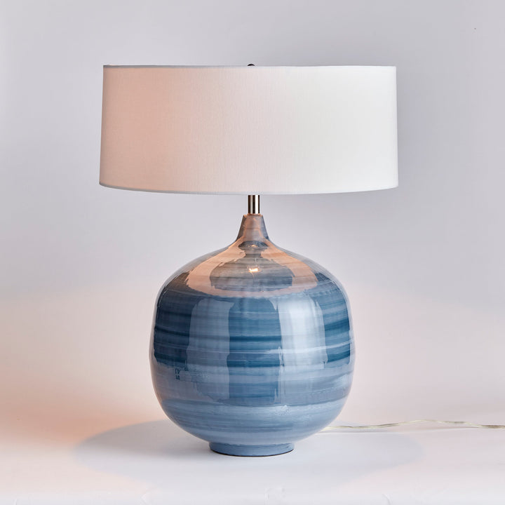 This blue table lamp features a unique design made of enameled iron and hand-painted for a lightweight yet elegant look. The horizontal swirl design adds a touch of refinement, while the oversized linen shade completes the modern aesthetic. A perfect addition to any contemporary living or dining space.