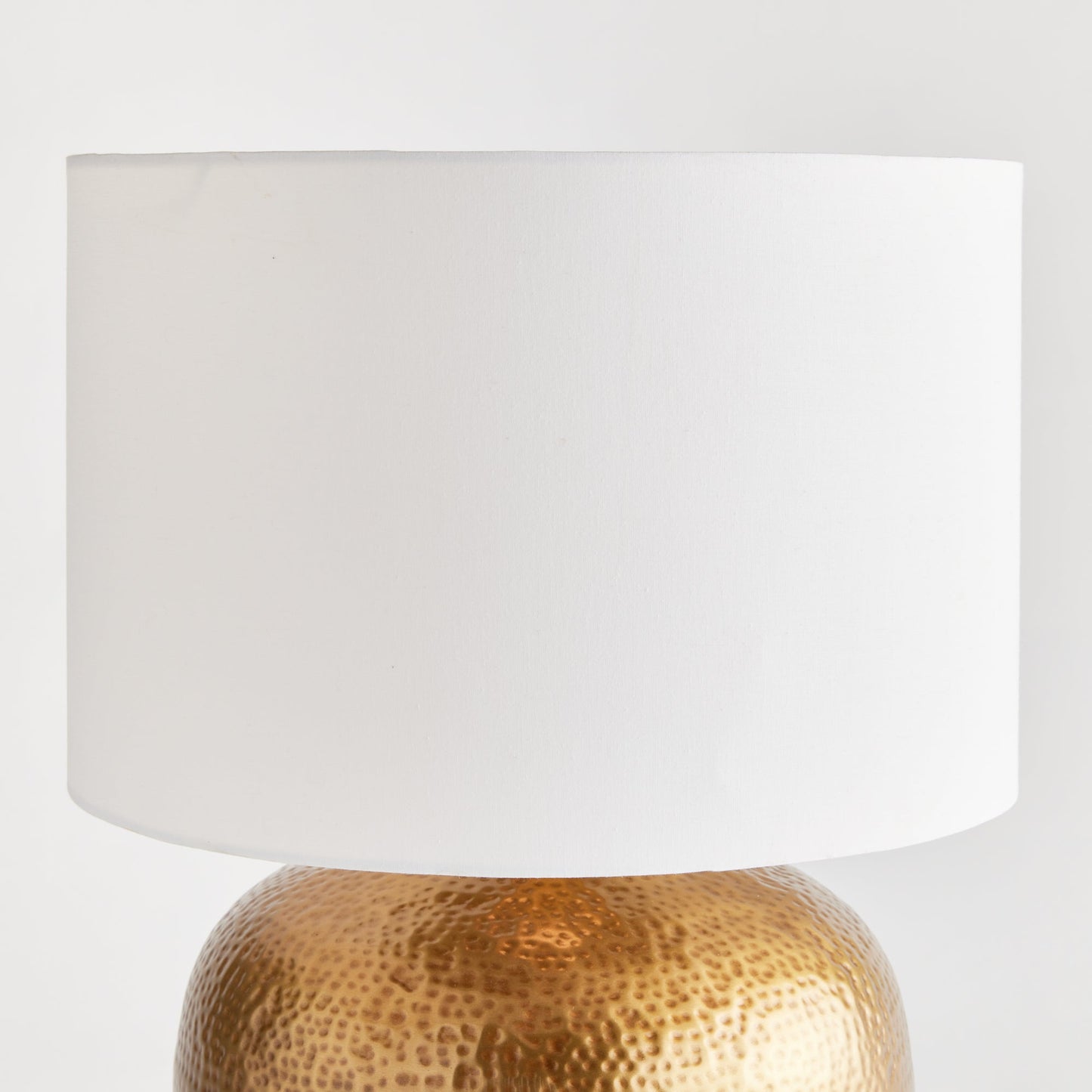 Crafted with a round base in warm brass and an impressive scale, the Tara Gold &amp; White Table Lamp serves as a handsome anchor for any room. Its clean white linen drum shade adds a soft touch and complements the bold design. Made from hammered iron, this one-of-a-kind lamp is sure to catch the eye and become a stunning focal point in your space.