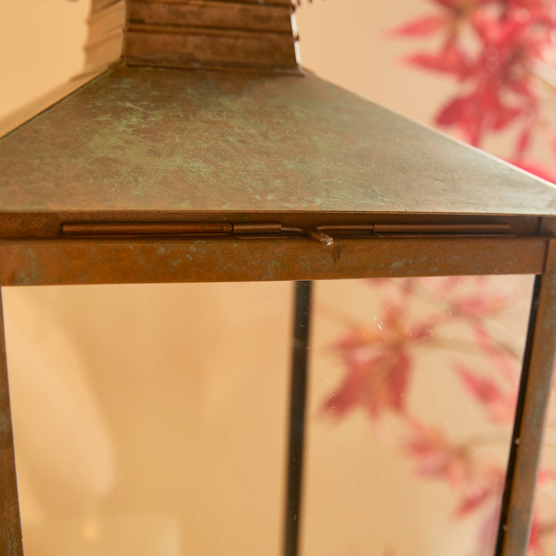 This lantern's design is characterized by a curved pagoda and a notched edge, adding to its simple yet distinctive look. The warm patina finish gives it a weathered appearance, making it a suitable choice for any indoor setting.