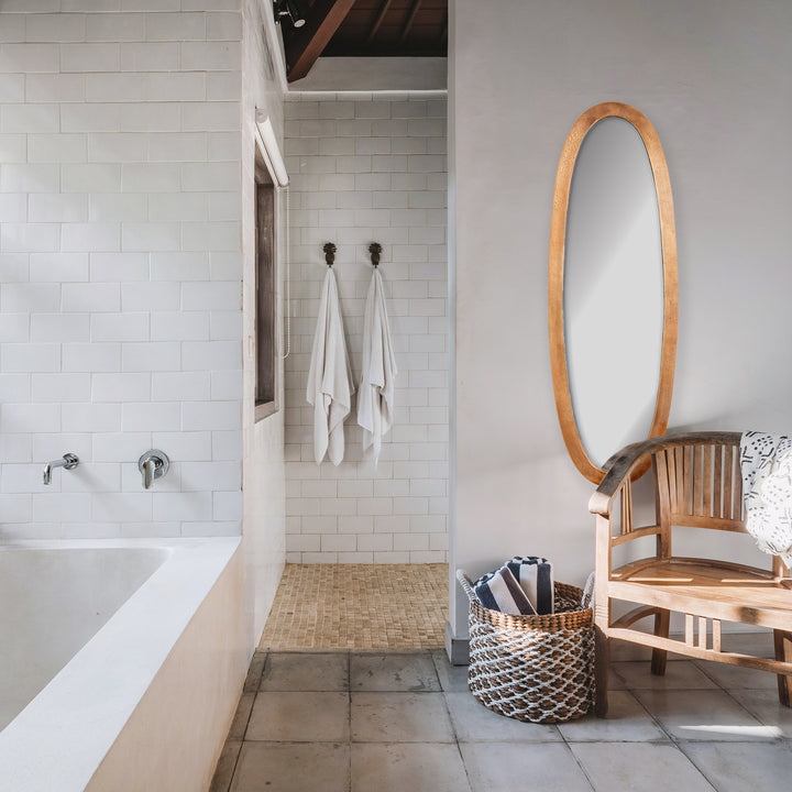 Ellery antique brass oval wall mirror hanging beside a wooden armchair and woven basket in a pale tiled room. Transitional