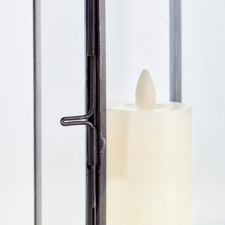 Detail of Emmett black lantern showing its side closure beside clear glass and an illuminated candle inside. Transitional