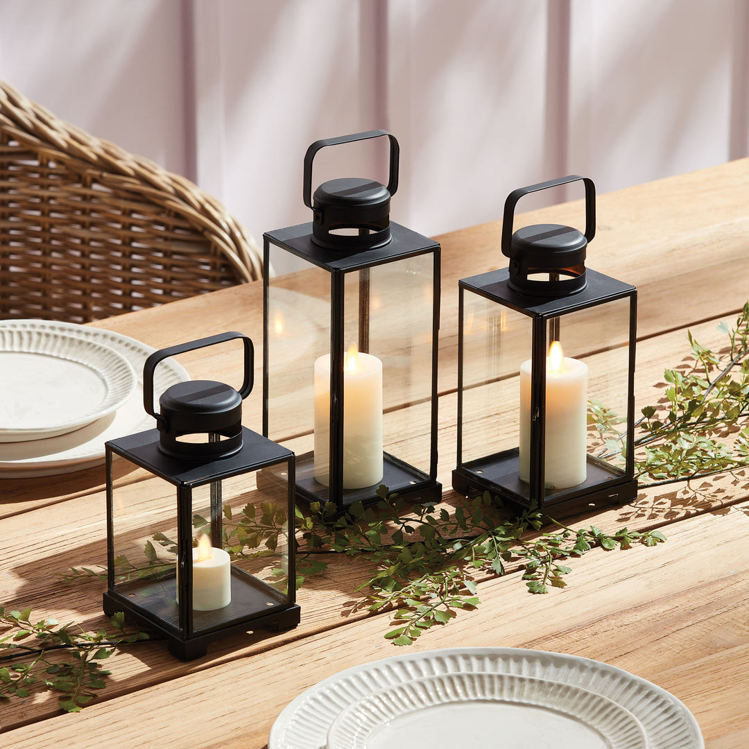 Emmett black glass lanterns in three sizes with candles, arranged along a wooden dining table beside greenery. Transitional