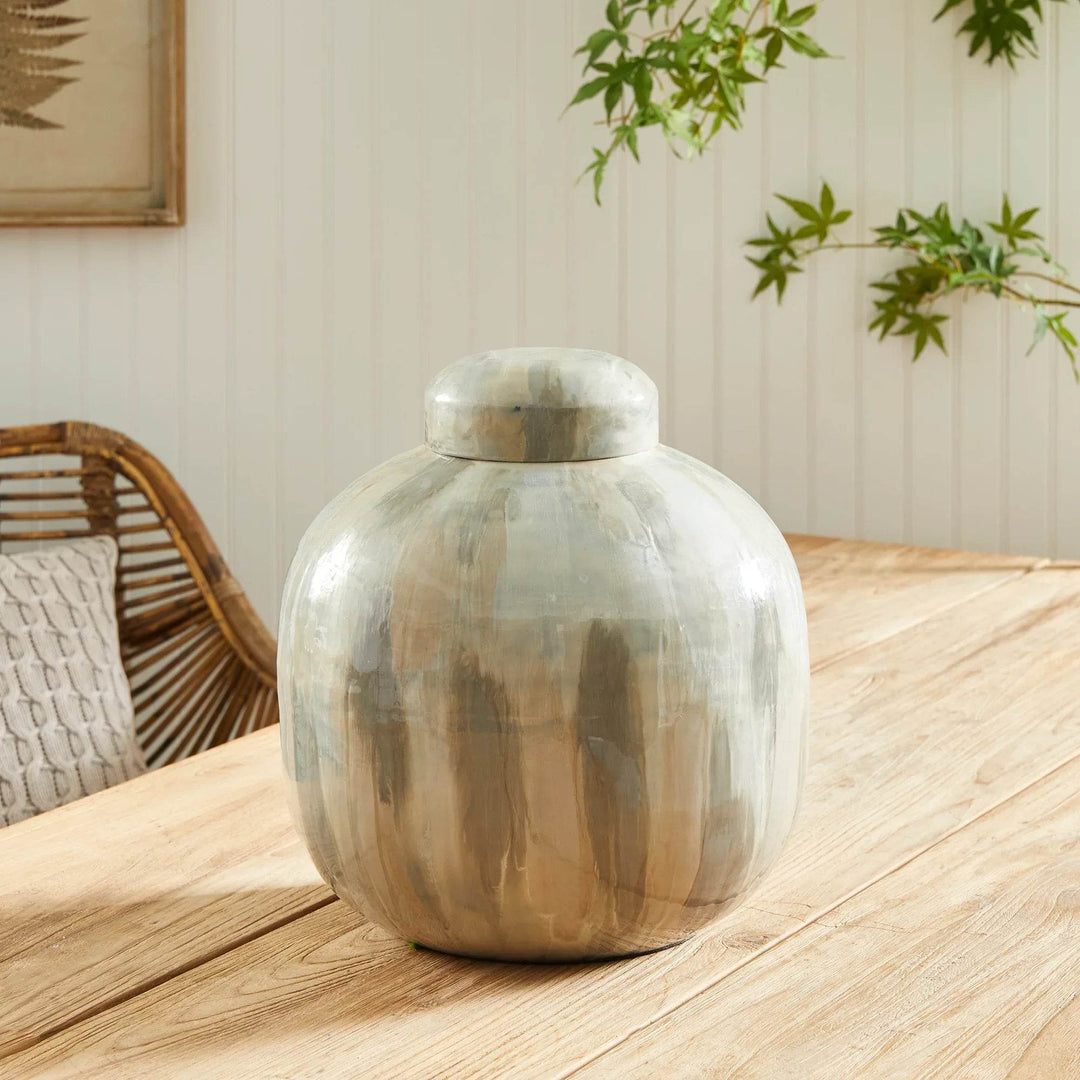 Enameled iron lidded urn with soft neutral brushwork and glossy sheen, styled on wood dining table with greenery in the background – Transitional