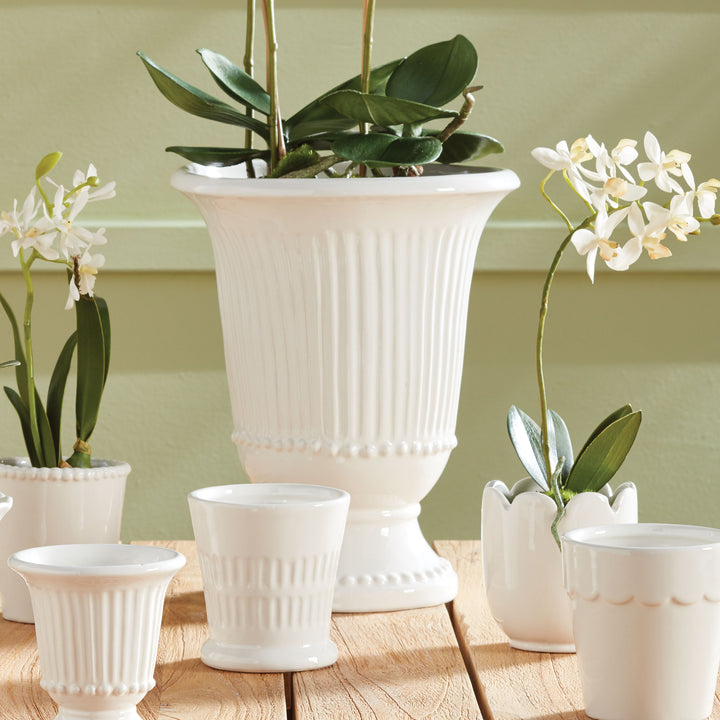 White fluted pedestal vase styled with white orchids among coordinating white vessels on a wide wooden table | Traditional