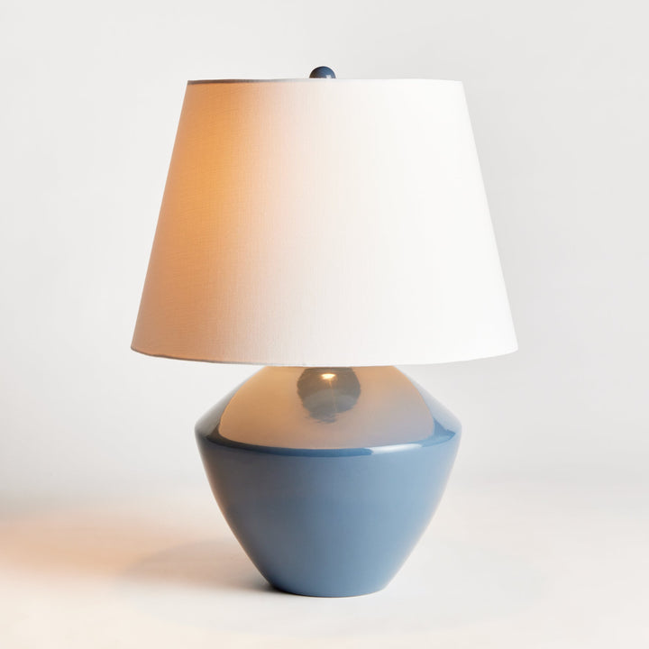 Crafted from a wooden mold dating back centuries, this stunning table lamp boasts a timeless silhouette and a deep blue finish. Topped with a tapered fabric shade, it adds a touch of history and elegance to any room, whether it be a family room, study, or bedroom. This ceramic blue table lamp is a dynamic addition to any space.