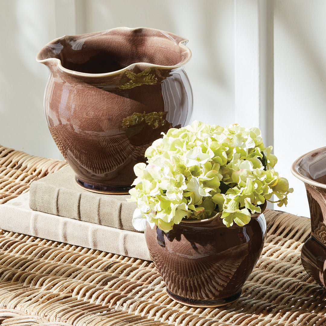 Brown ceramic planter set styled on a woven table with flowers in the smaller pot and the larger pot empty. Transitional