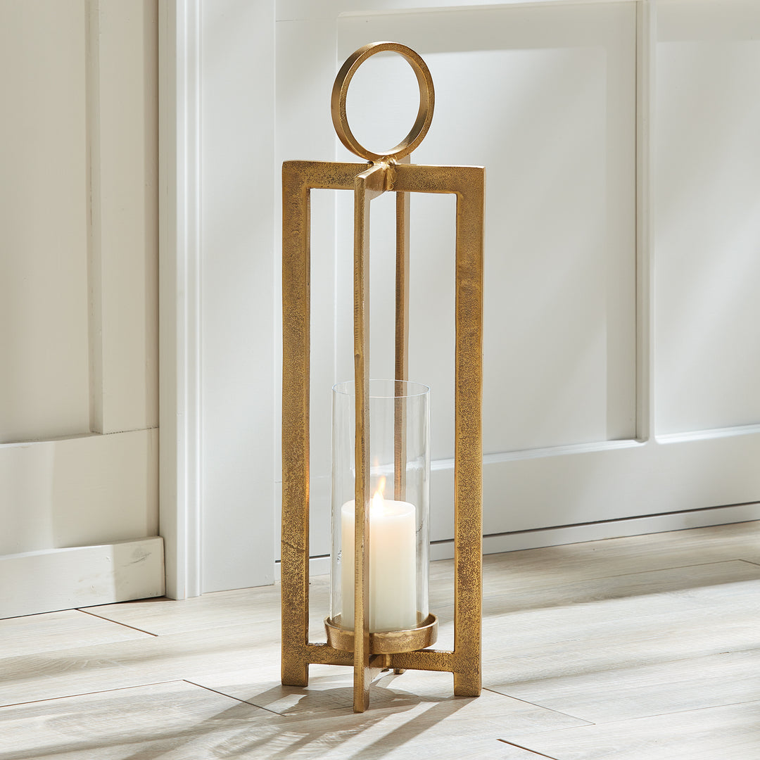 Large antique gold geometric metal lantern with glass hurricane styled on floor in bright interior beside wall paneling, Transitional style
