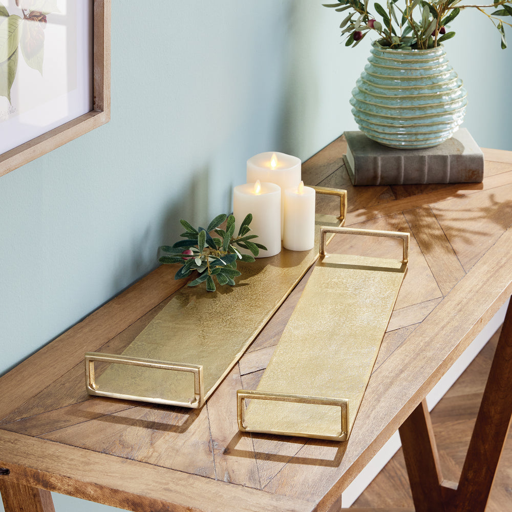 Two narrow gold decorative trays styled with ivory candles and greenery on a long natural wood console table – Transitional.