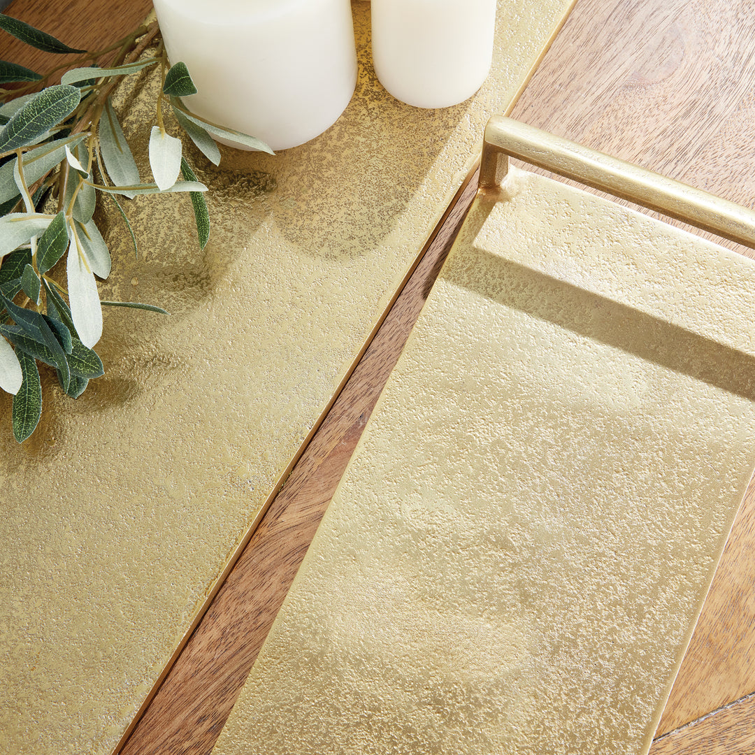 Gold narrow decorative trays styled with ivory pillar candles and leafy greenery on a warm wood tabletop – Transitional.