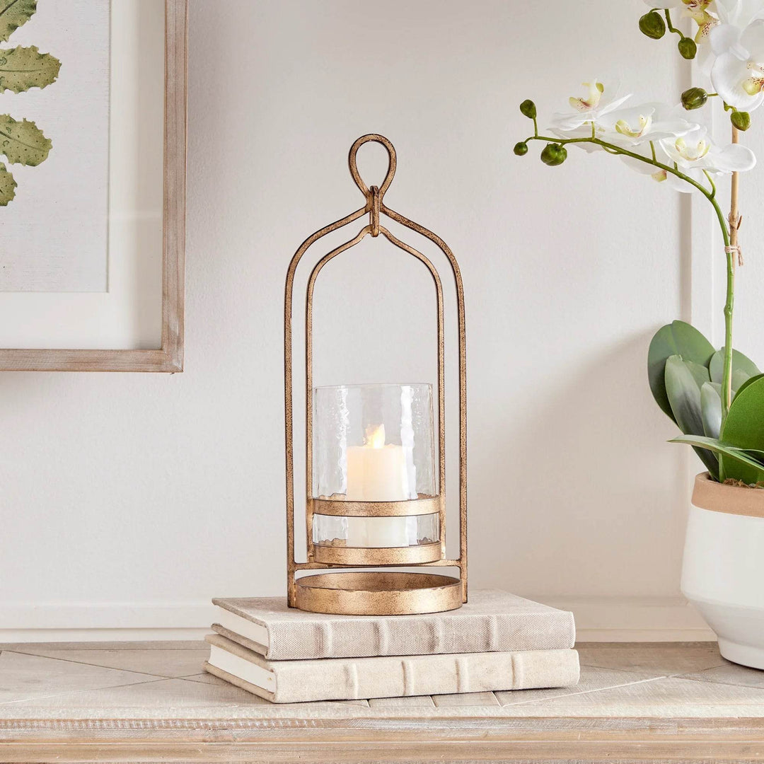 Small gold hurricane candle holder with hammered glass cylinder styled on books and console table, antique iron frame with loop handle – Transitional
