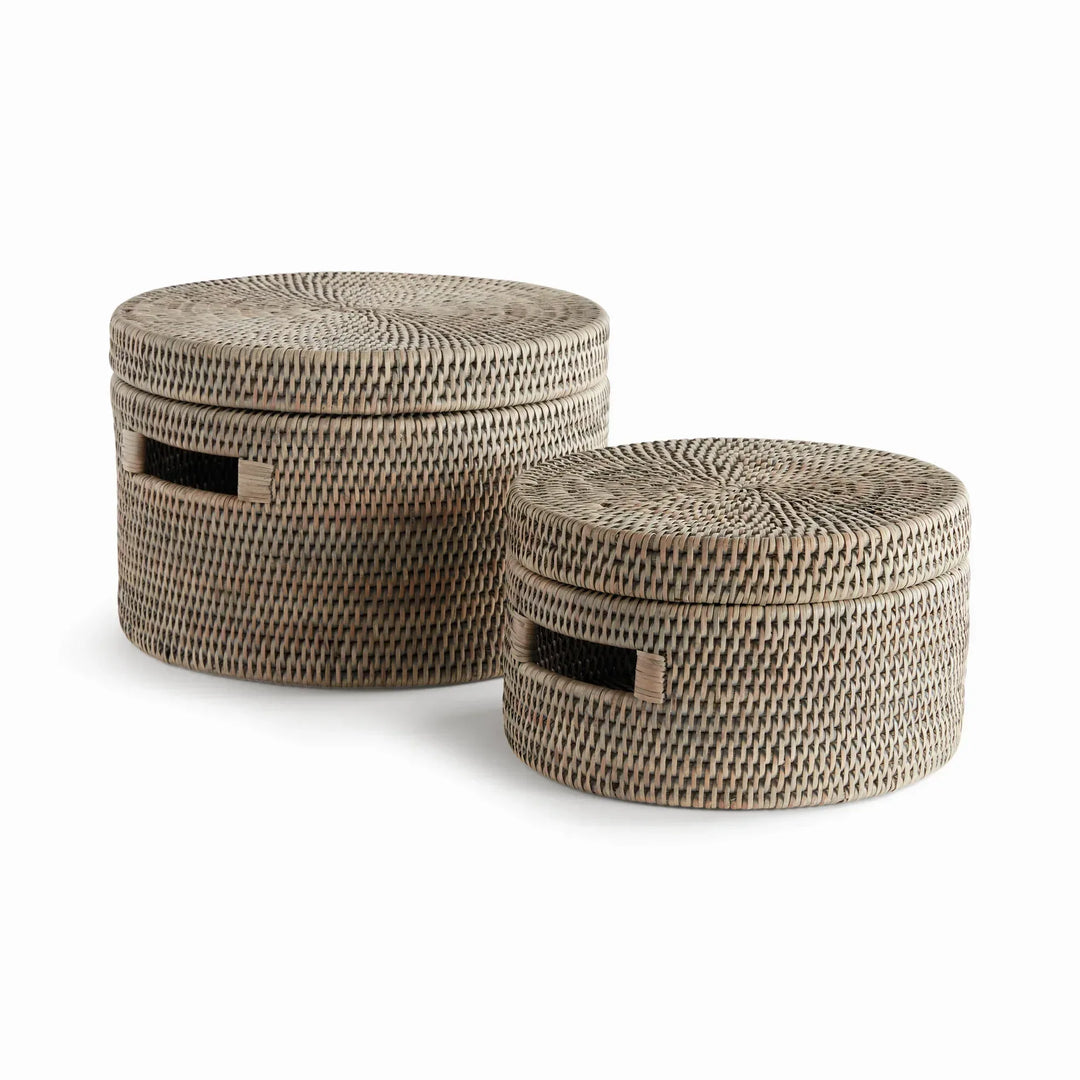 Two graywash handwoven rattan round lidded boxes with cutout handles, shown as a graduated set against white, Transitional