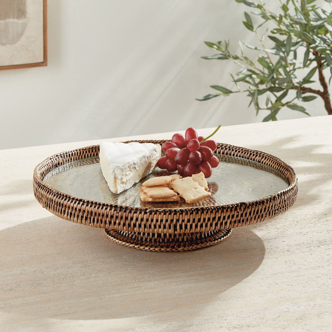 Hadley warm brown rattan stand with glass insert styled with grapes, cheese and crackers on a wood table by foliage. Coastal