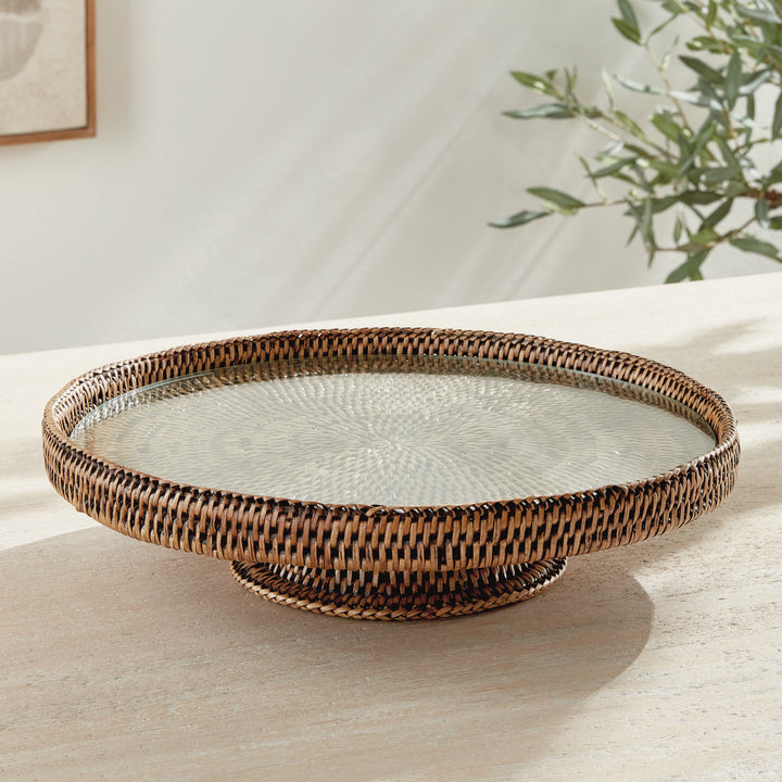 Hadley warm brown rattan pedestal stand with glass insert on a pale wood table in a softly lit interior setting. Coastal