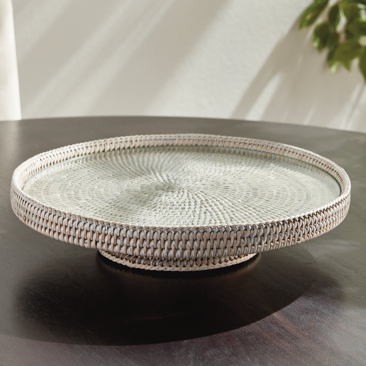 Hadley whitewash round rattan pedestal stand with clear glass insert on a dark wood table in a lit interior setting. Coastal