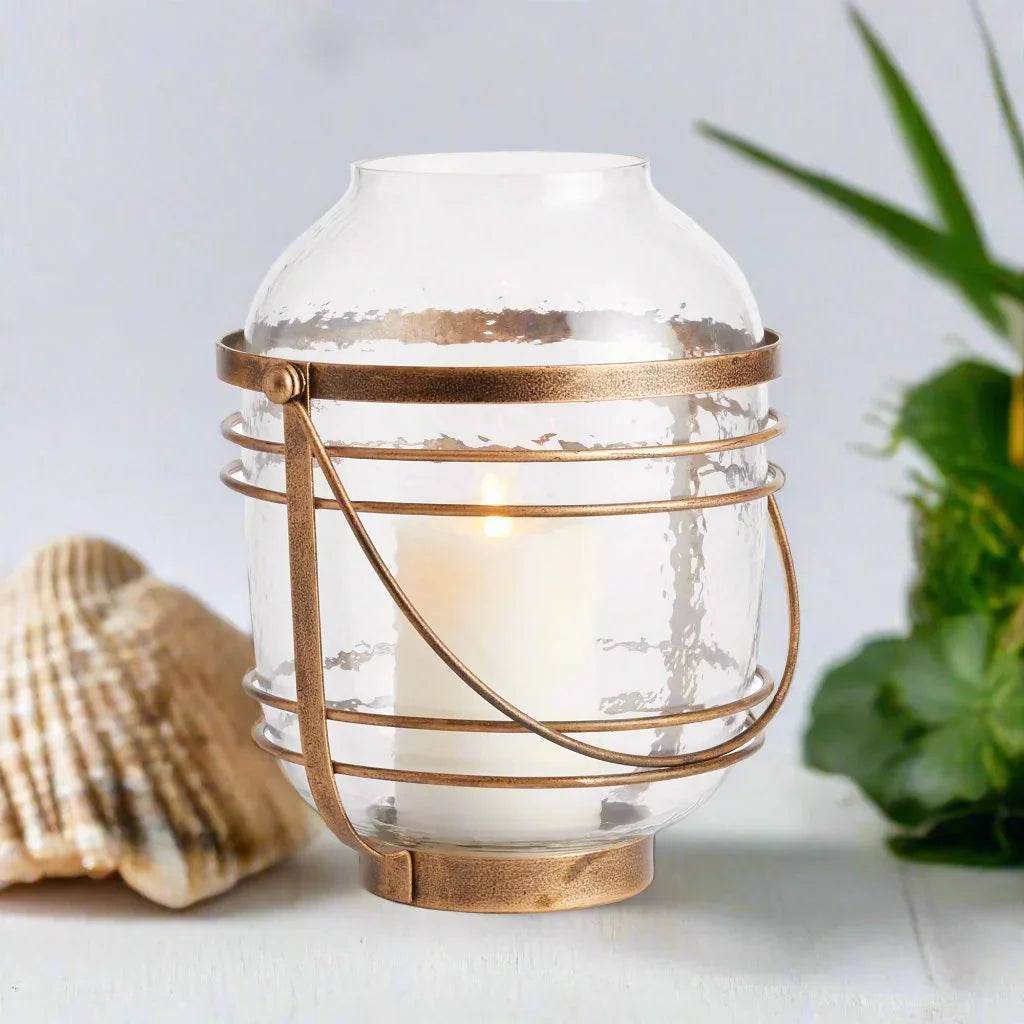Large hammered glass hurricane with antique gold iron bands and base, styled on tabletop with seashell for glow – Coastal
