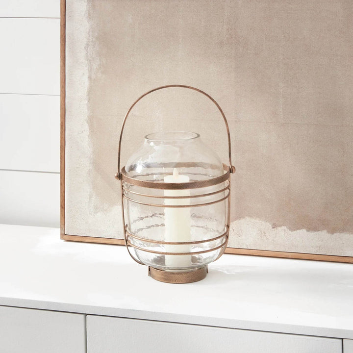 Hammered glass hurricane with antique gold iron band and handle, shown on shelf against artwork for soft light – Coastal