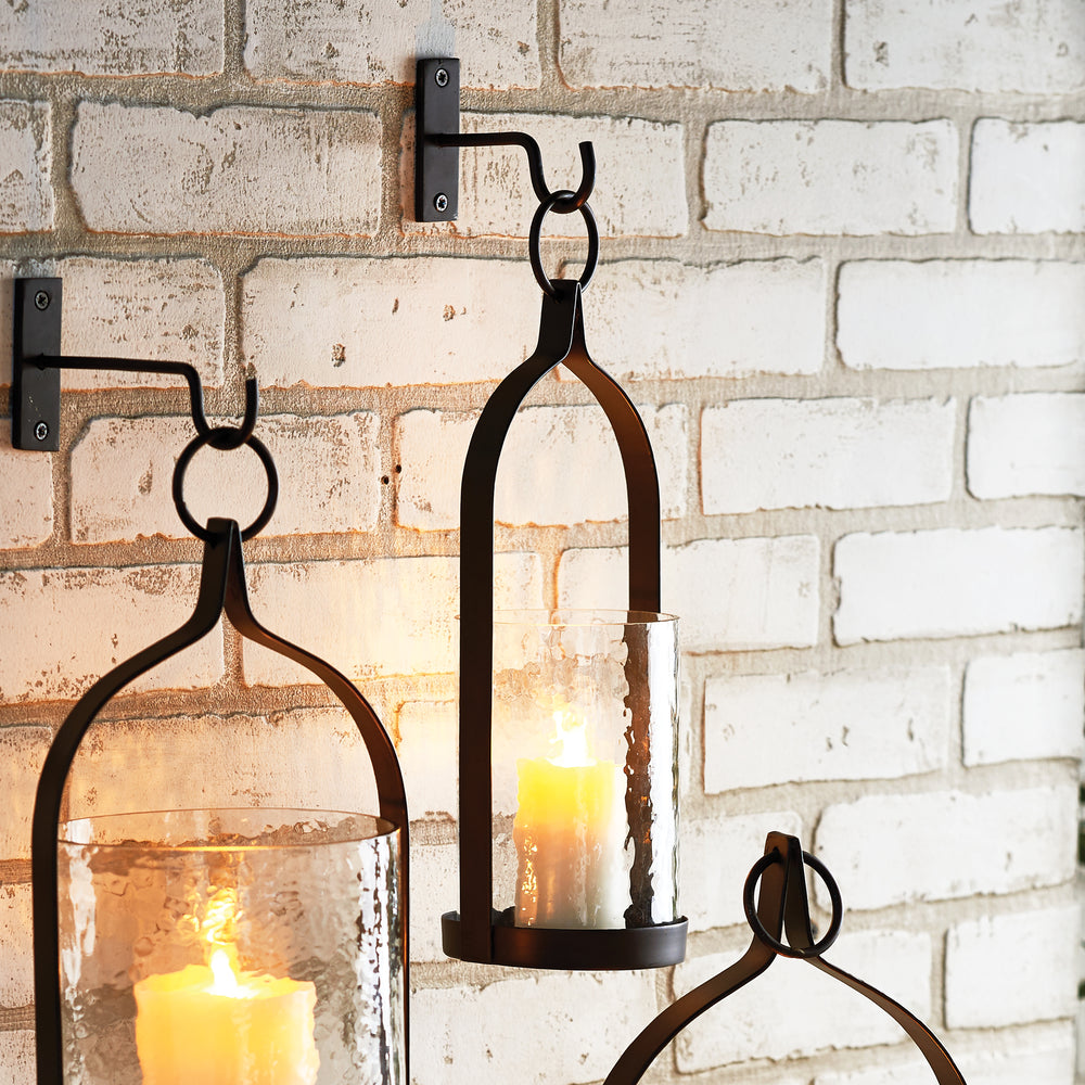 Harlan iron lanterns in graduated sizes hanging from hooks against pale brick, with illuminated candles inside. Transitional