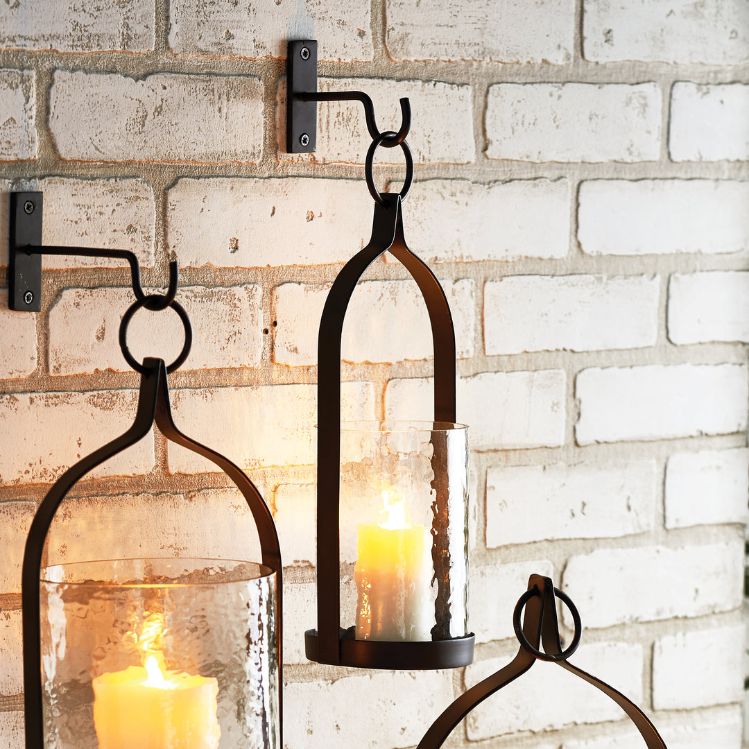 Harlan iron lanterns in graduated sizes hanging from hooks against pale brick, with illuminated candles inside. Transitional