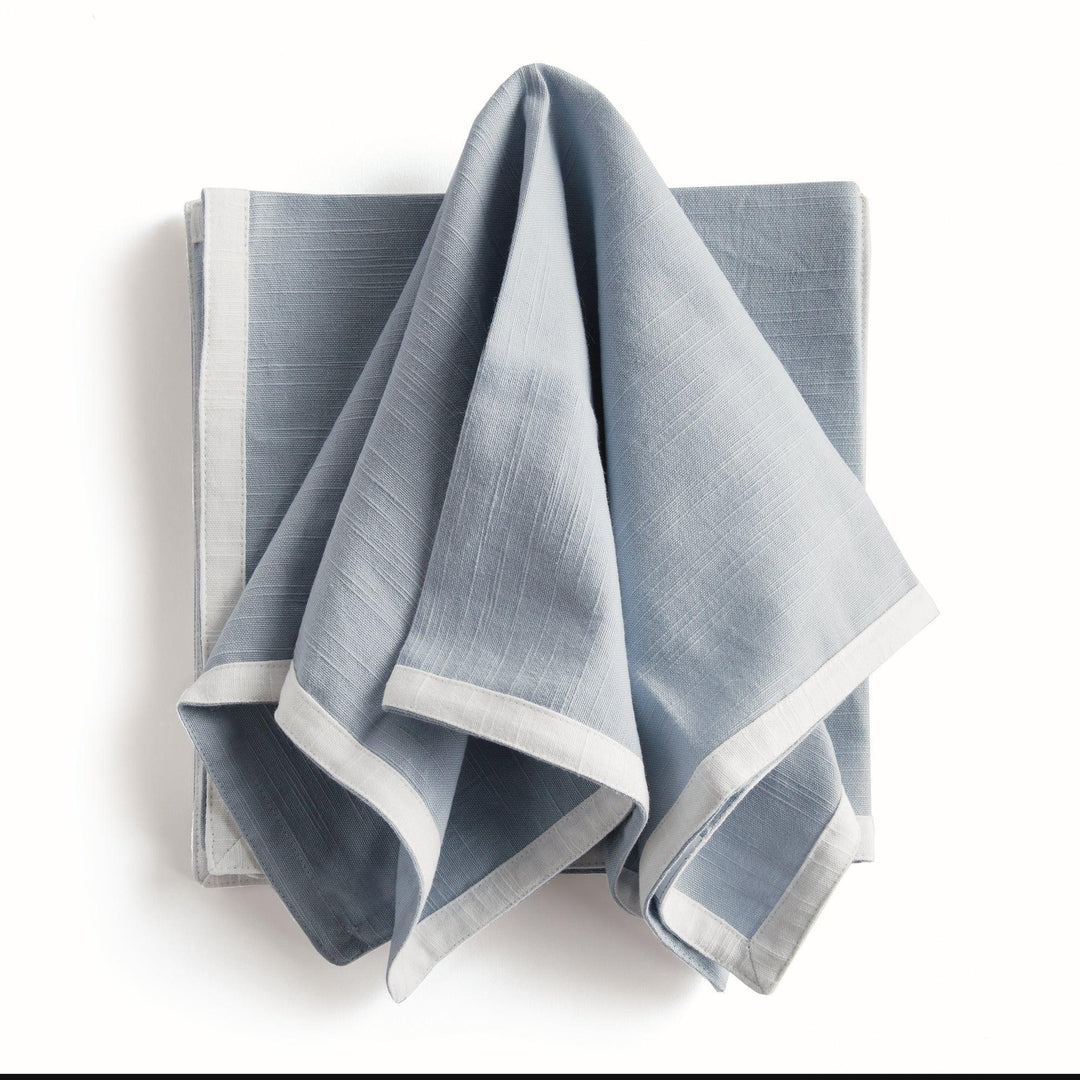 Blue and white dinner napkins, draped over a folded stack, showing the contrast border and the cotton texture. Transitional