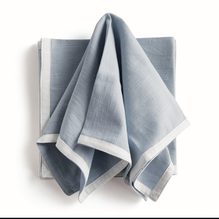Blue and white dinner napkins, draped over a folded stack, showing the contrast border and the cotton texture. Transitional