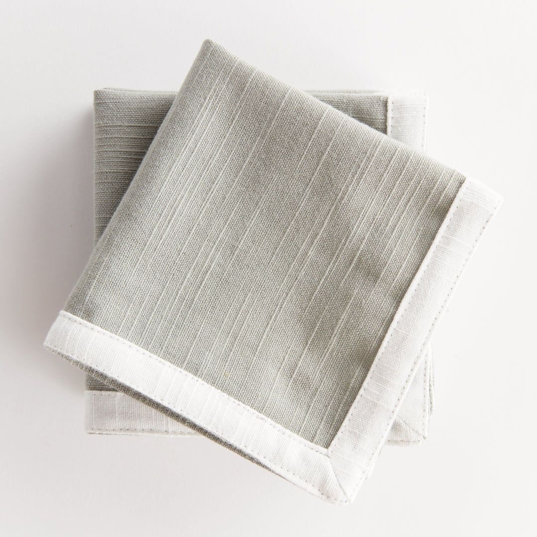 Green and white cocktail napkins, layered and folded, showing green cotton panels framed by white cloth borders. Transitional