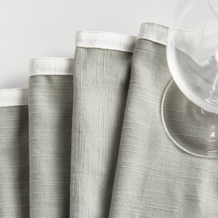 Green and white cocktail napkins, folded beside glasses, showing the white border and soft green cotton surface. Transitional