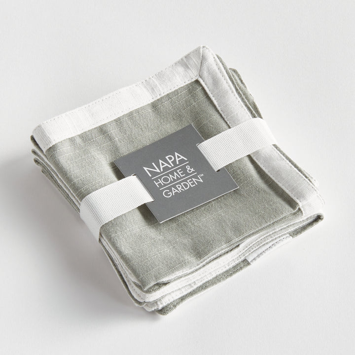 Green and white cocktail napkins, folded as a set tied with ribbon, with white borders around the cotton fabric. Transitional