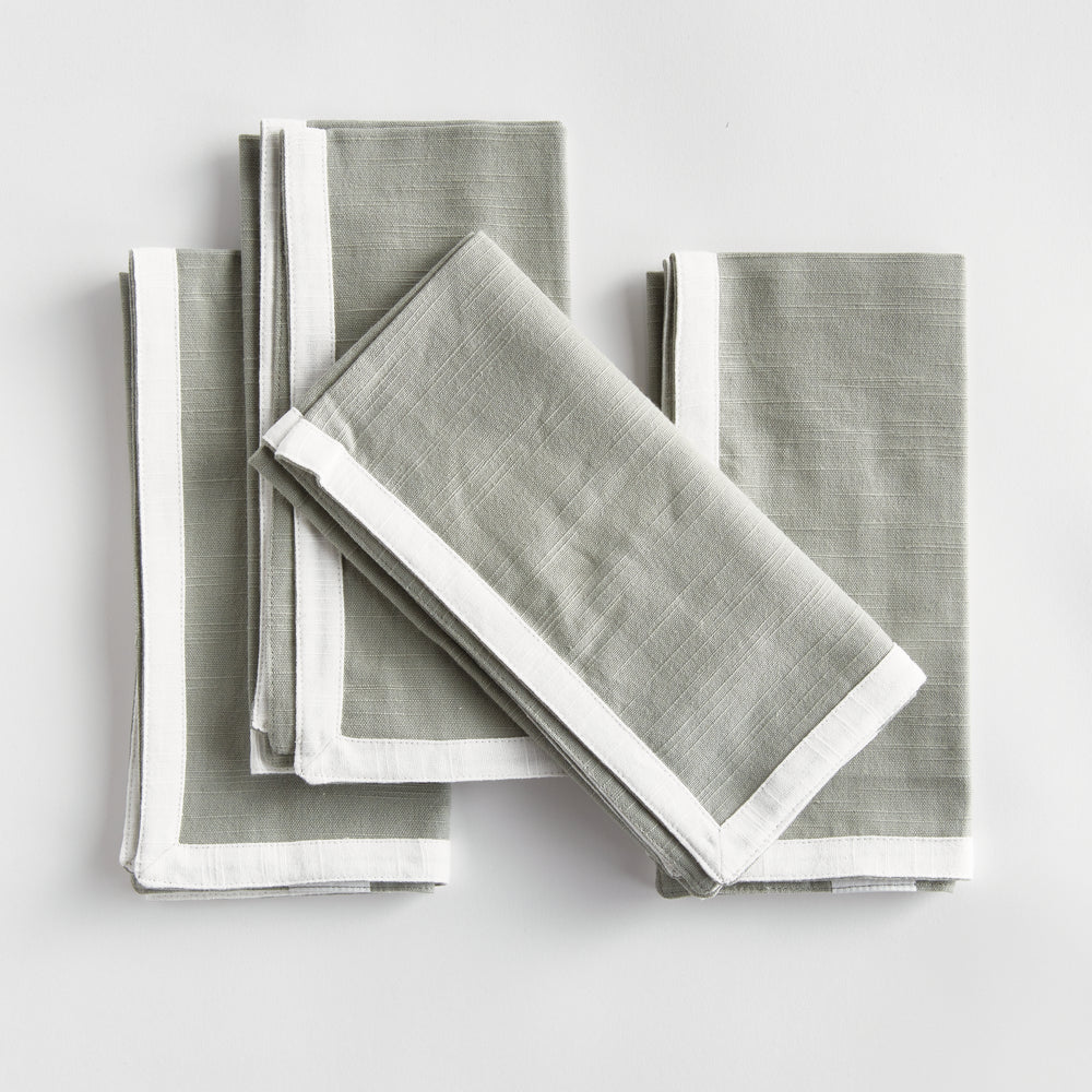 Green and white dinner napkins, folded in a layered arrangement, showing the border and square cotton fabric. Transitional