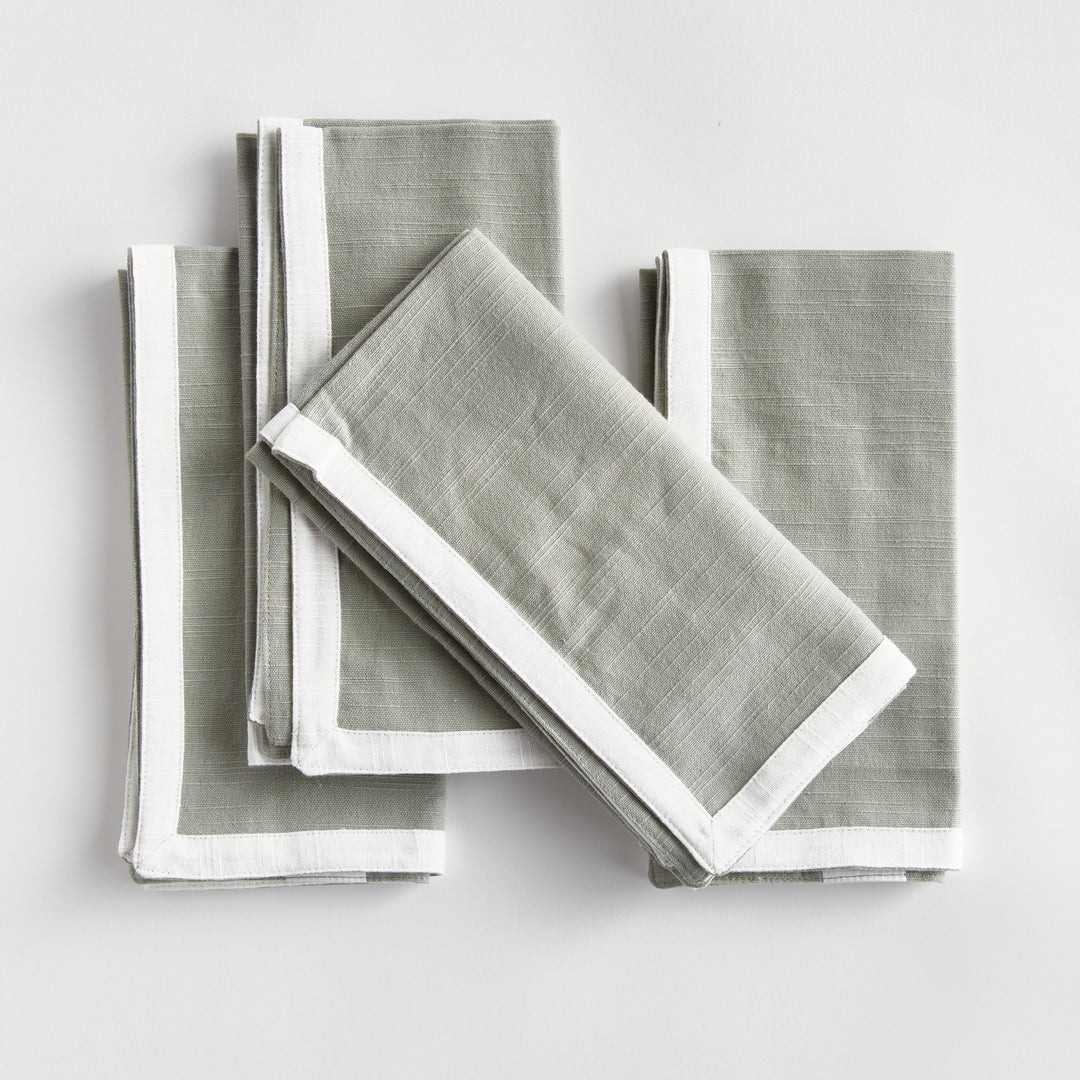 Green and white dinner napkins, folded in a layered arrangement, showing the border and square cotton fabric. Transitional
