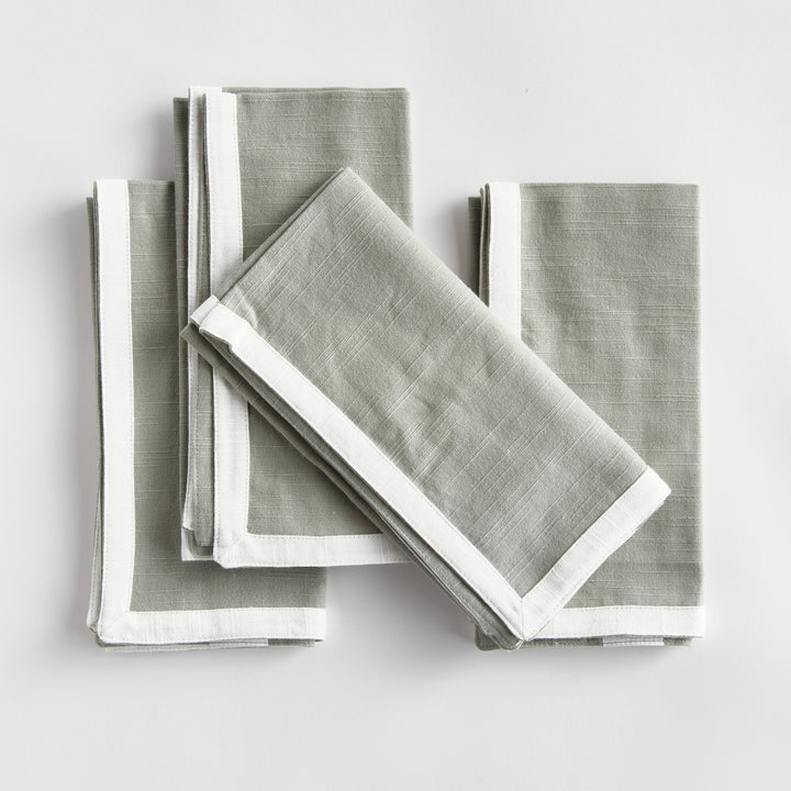 Green and white dinner napkins, folded in a layered arrangement, showing the border and square cotton fabric. Transitional