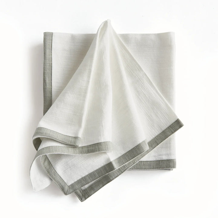 White and sage dinner napkins, draped over a folded stack, showing the contrast border and the cotton texture. Transitional