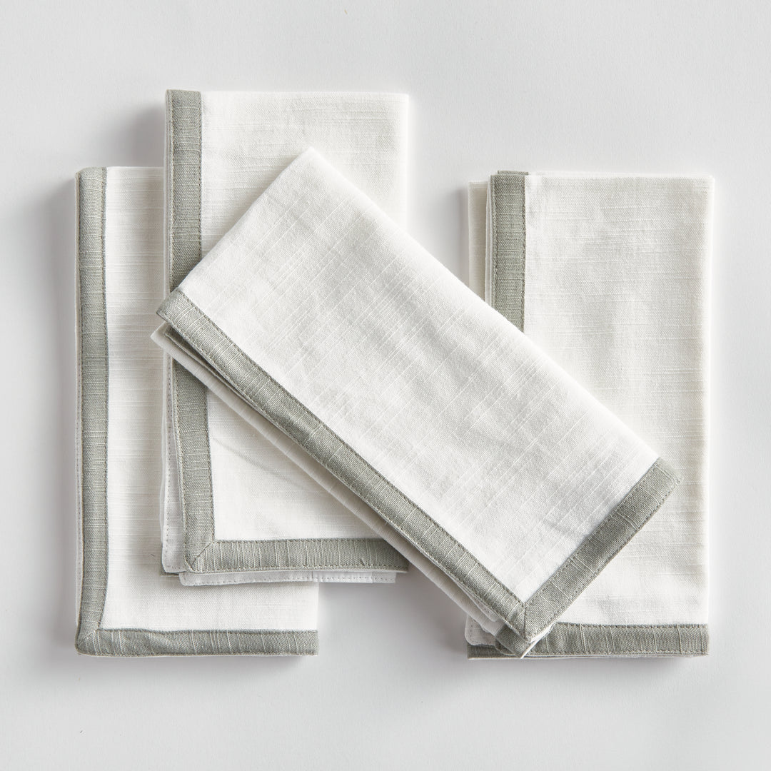 White and sage dinner napkins, folded in a layered arrangement, showing the border and square cotton fabric. Transitional
